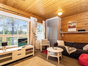 Interior acogedor de una cabaña con techo de madera, un televisor sobre un soporte blanco, un armario gris, un sofá y un sillón de peluche rojo.