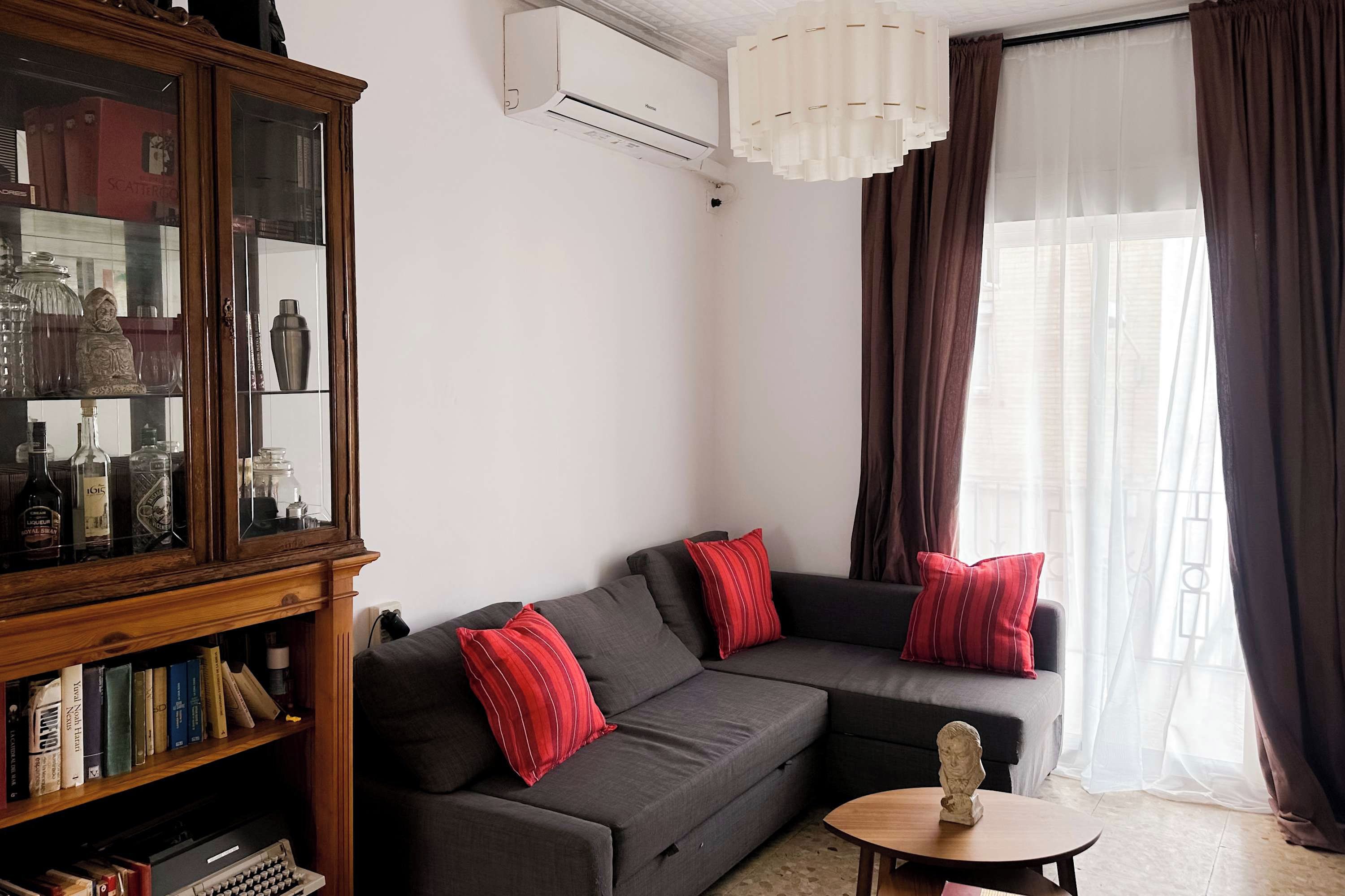 Un salón con un sofá gris oscuro, almohadas rojas a rayas, un aparador de madera con puertas de cristal y una pequeña mesa redonda con una escultura de busto.