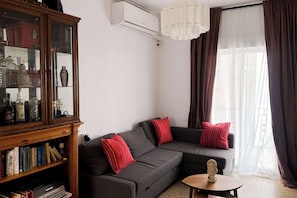 Een woonkamer met een donkergrijze bank, rode gestreepte kussens, een houten kast met glazen deuren en een kleine ronde tafel met een buste beeldhouwwerk.