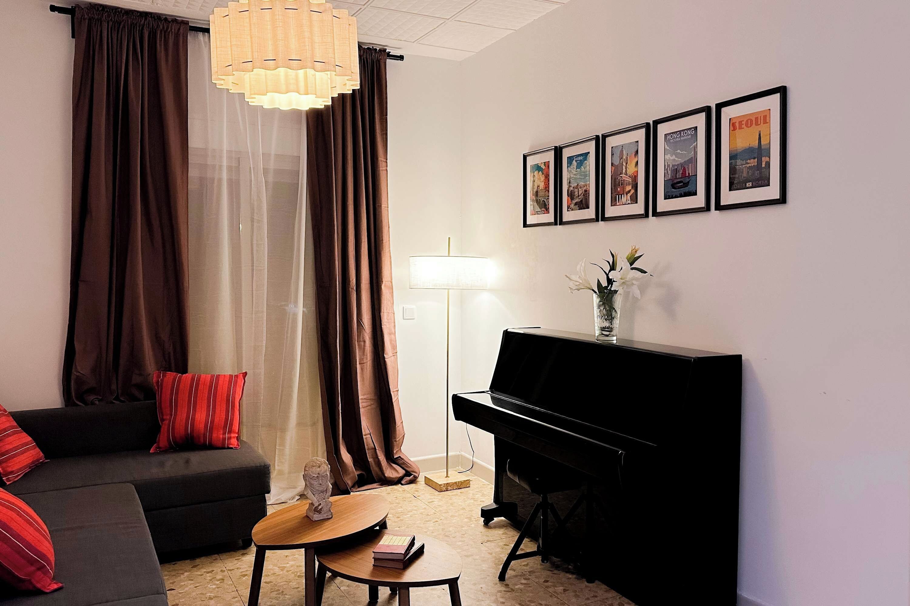 Un salon avec un piano noir, un canapé avec des coussins rouges, un grand lampadaire et un mur orné de tableaux encadrés.