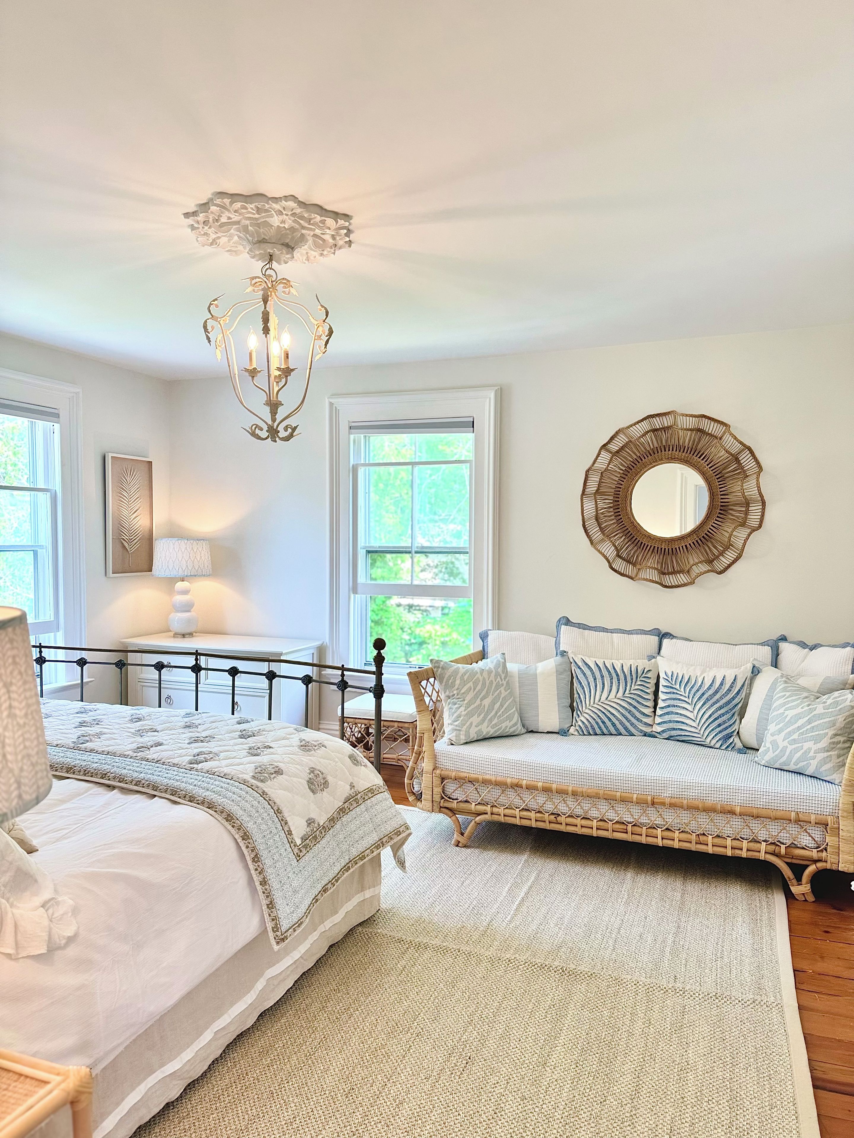 A bedroom with a bed, a wicker sofa, a chandelier, a mirror, and a window with a view of greenery.
