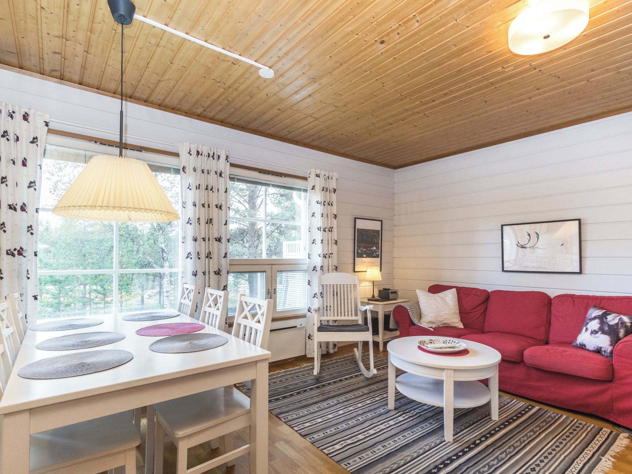 Una habitación acogedora con techo de madera, un sofá rojo, una mesa blanca con individuales y una ventana con cortinas.