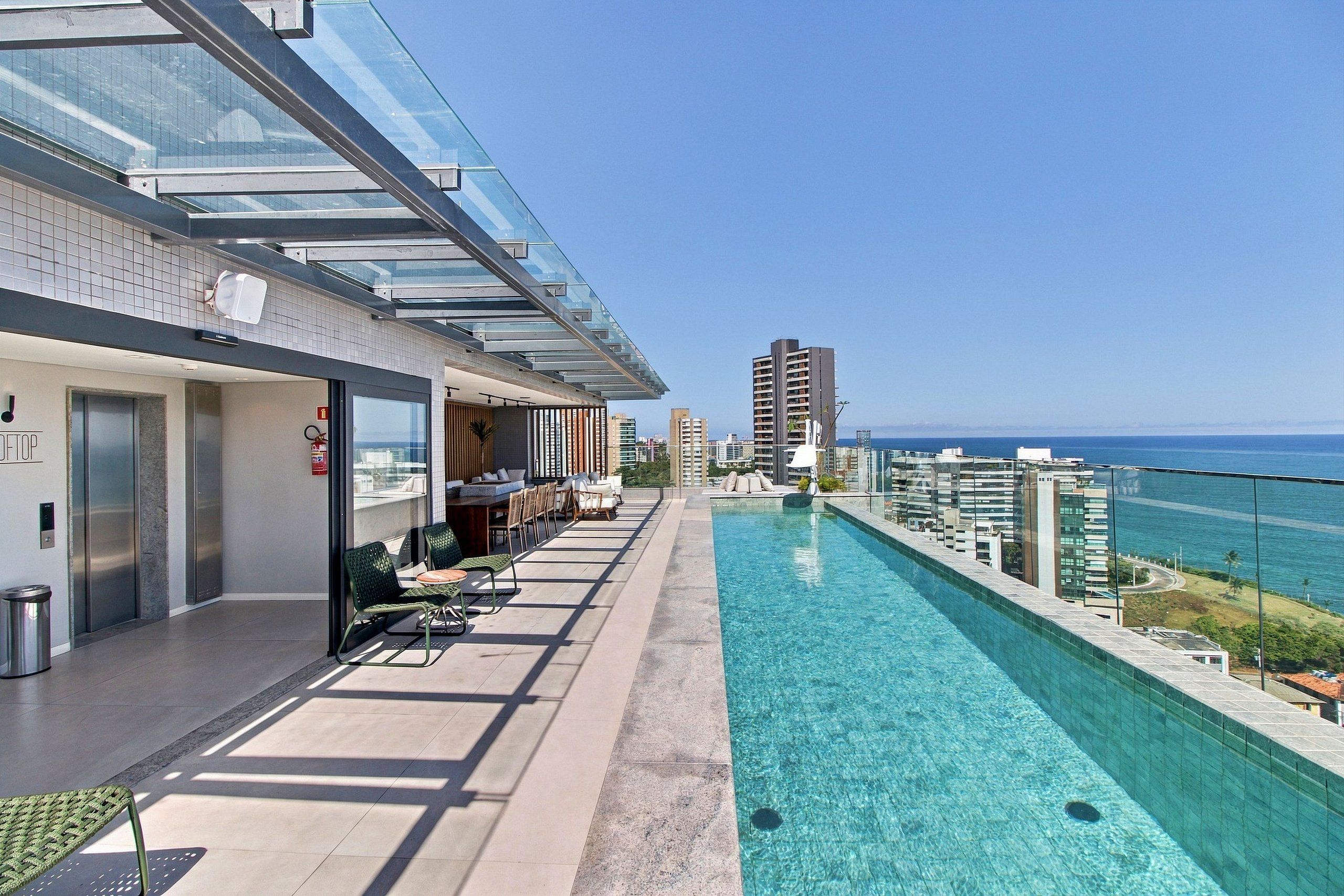 Ein Pool auf dem Dach mit Glasbrüstung, Sitzgelegenheiten im Freien und Blick auf Stadt und Meer.