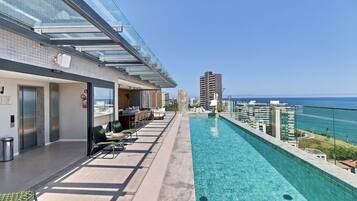 Ein Pool auf dem Dach mit Glasbrüstung, Sitzgelegenheiten im Freien und Blick auf Stadt und Meer.