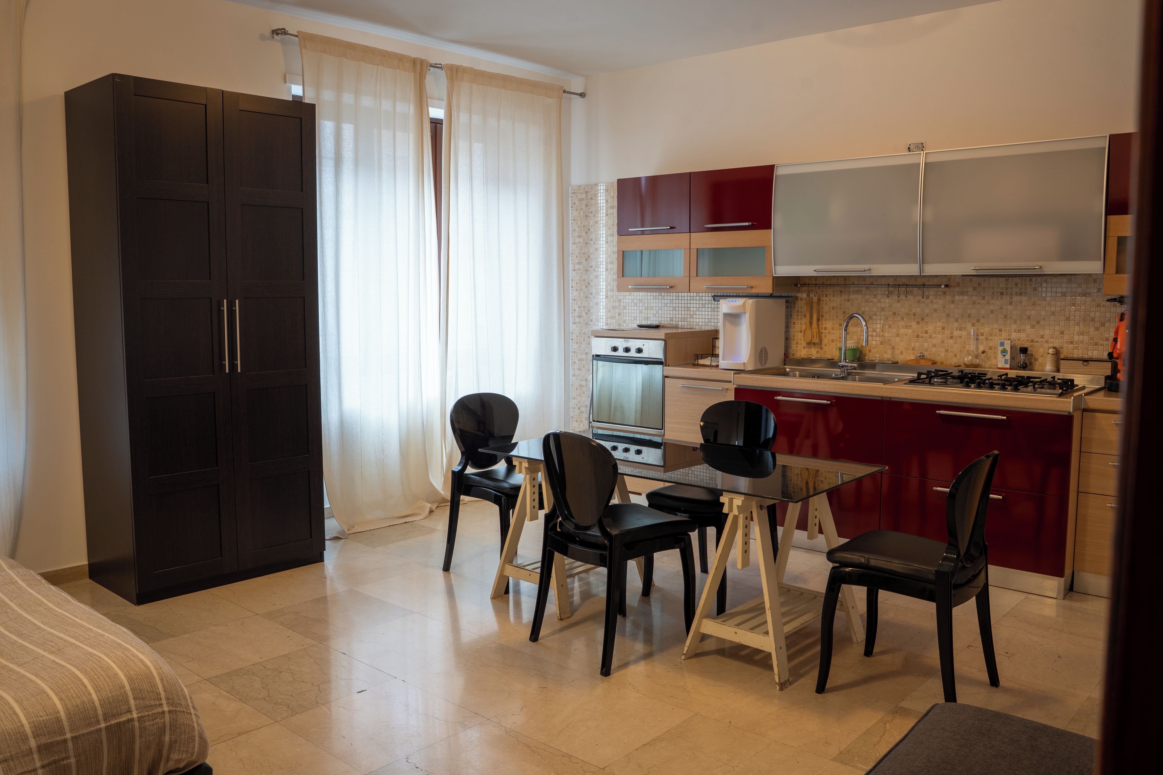 A compact kitchen with a dining area, a dark wardrobe, and a window with curtains.