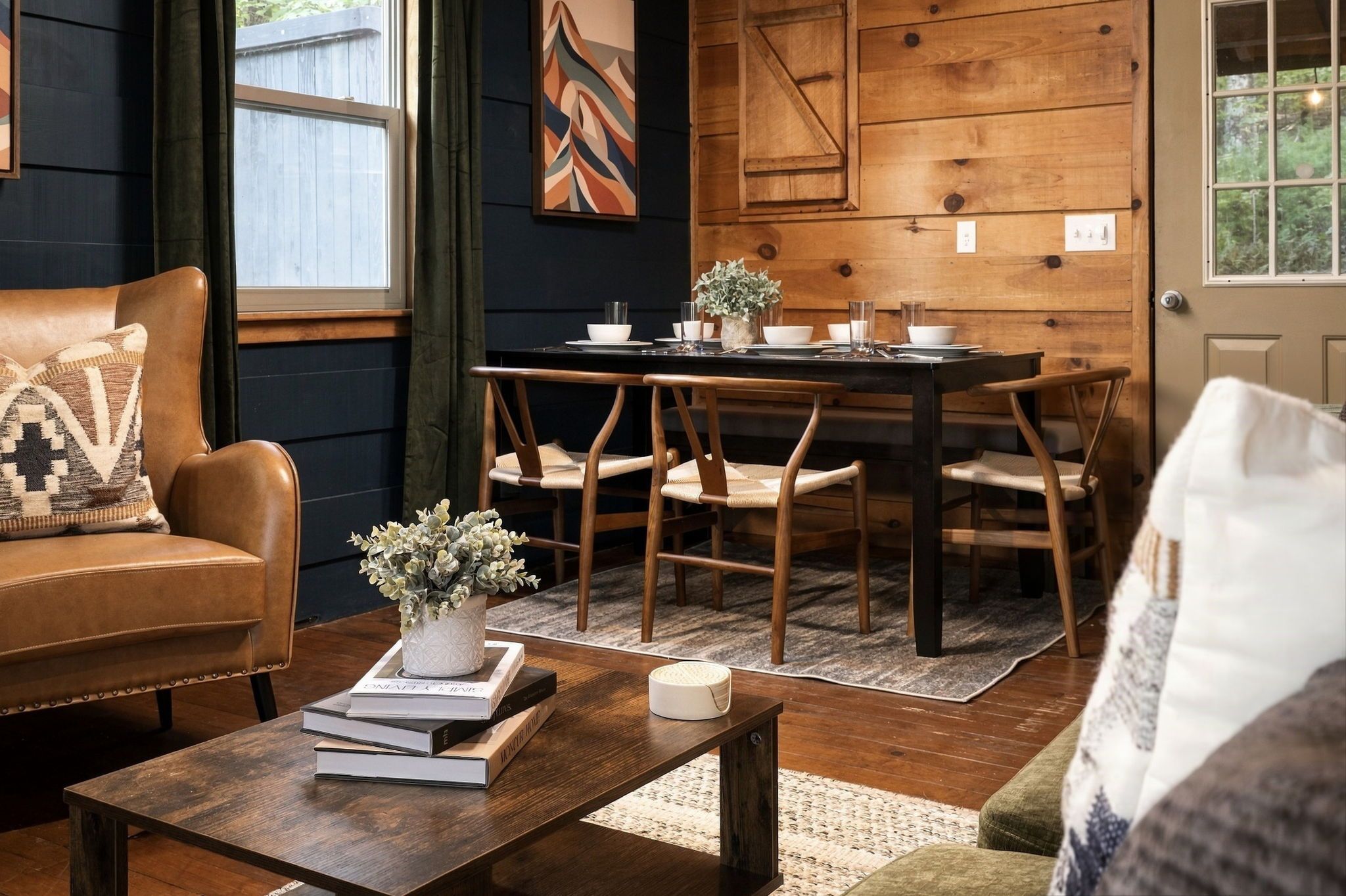 Un salon chaleureux avec un canapé en cuir, une table basse en bois agrémentée de livres et d’un vase, un coin repas avec une table et des chaises en bois, et une porte ornée d’un panneau mural aux motifs géométriques.