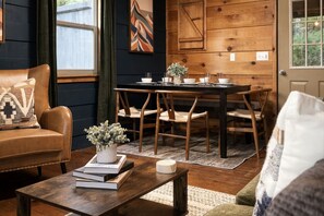A cozy living room with a leather sofa, a wooden coffee table with books and a vase, a dining area with a wooden table and chairs, and a door with a geometric wall panel.