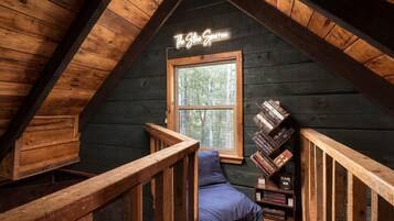 Une pièce mansardée chaleureuse avec un plafond en pente, une fenêtre donnant sur les arbres, un panneau néon sur lequel on peut lire « The Store Sparrow », et une bibliothèque remplie de livres.
