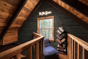 A cozy attic room with a sloped ceiling, a window with a view of trees, a neon sign reading 'The Store Sparrow', and a bookshelf filled with books.