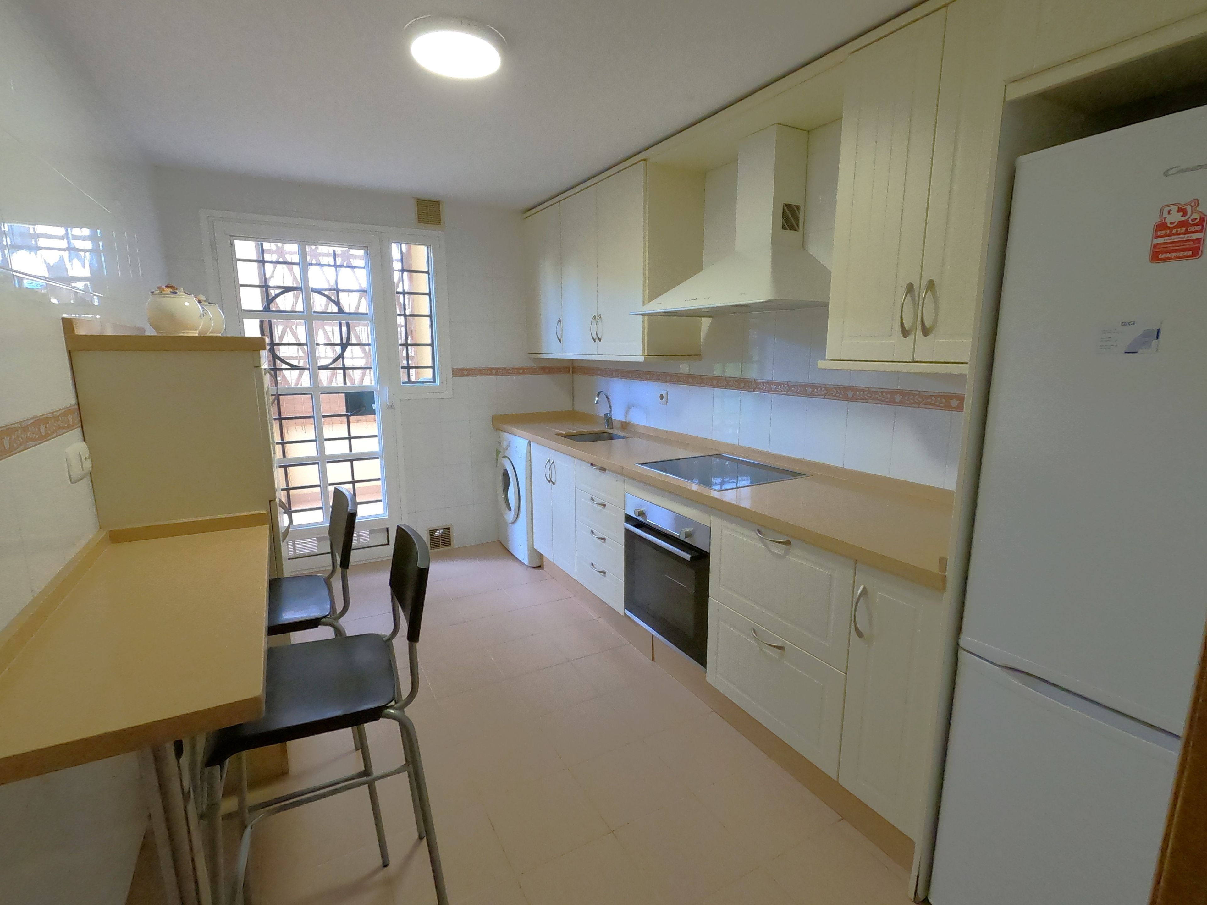 Une cuisine de taille réduite, équipée d’un évier, d’une cuisinière et d’un réfrigérateur. Une fenêtre avec des barreaux et une porte donnant sur un espace extérieur sont également présents.