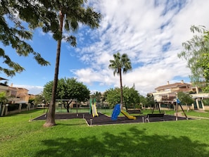 A park with playground equipment, including slides and swings, surrounded by grass and trees. - Apartment 'Sunny Terrace by the Golf Course' with Private Terrace, Wi-Fi and Air Conditioning (Vélez-Málaga)