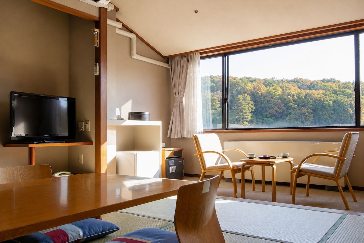 A modern room with a flat-screen TV, a wooden dining table, chairs, a small kitchen area, and a large window offering a view of a forest.