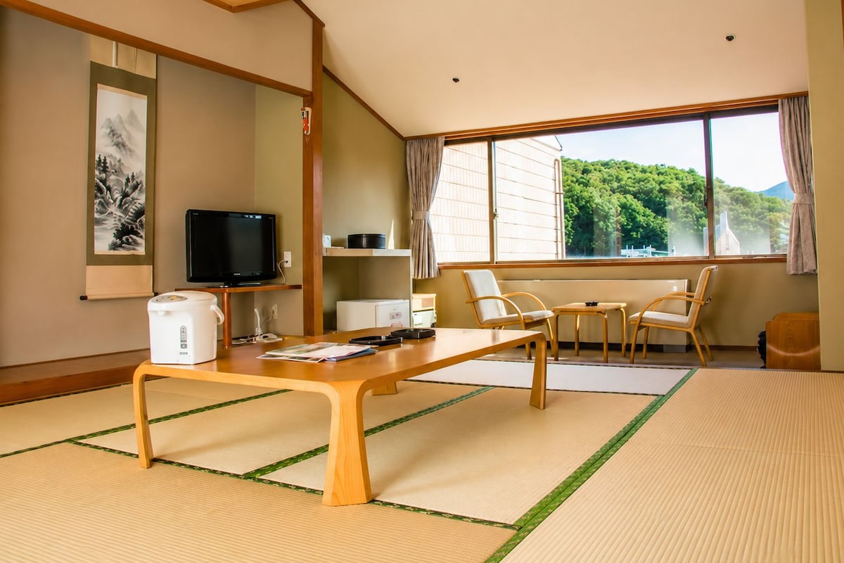 A traditional Japanese-style room with tatami flooring, a low table, a television, and a view of greenery outside.