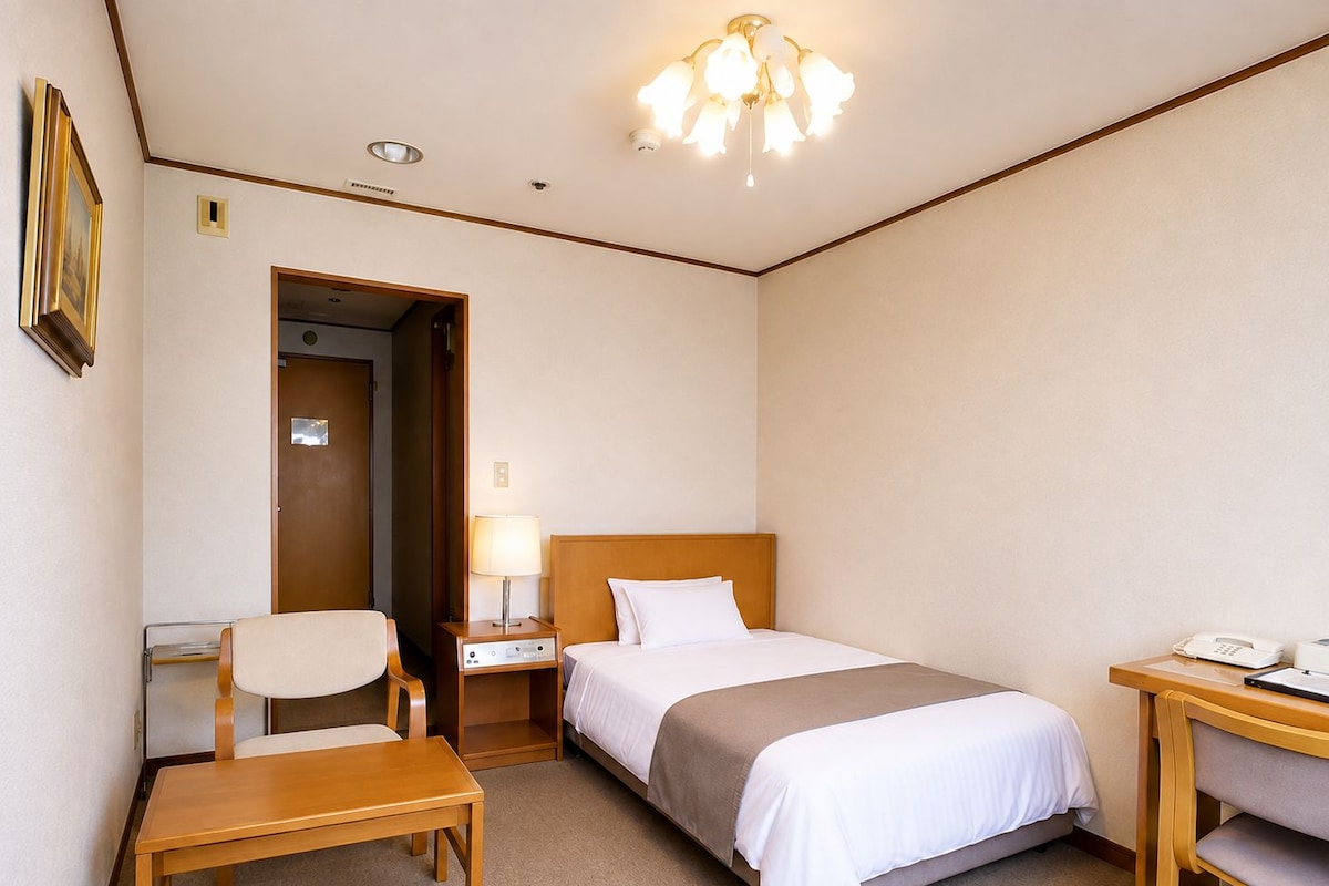 A hotel room with a bed, bedside table, chair, desk, lamp, and a door leading to another room.