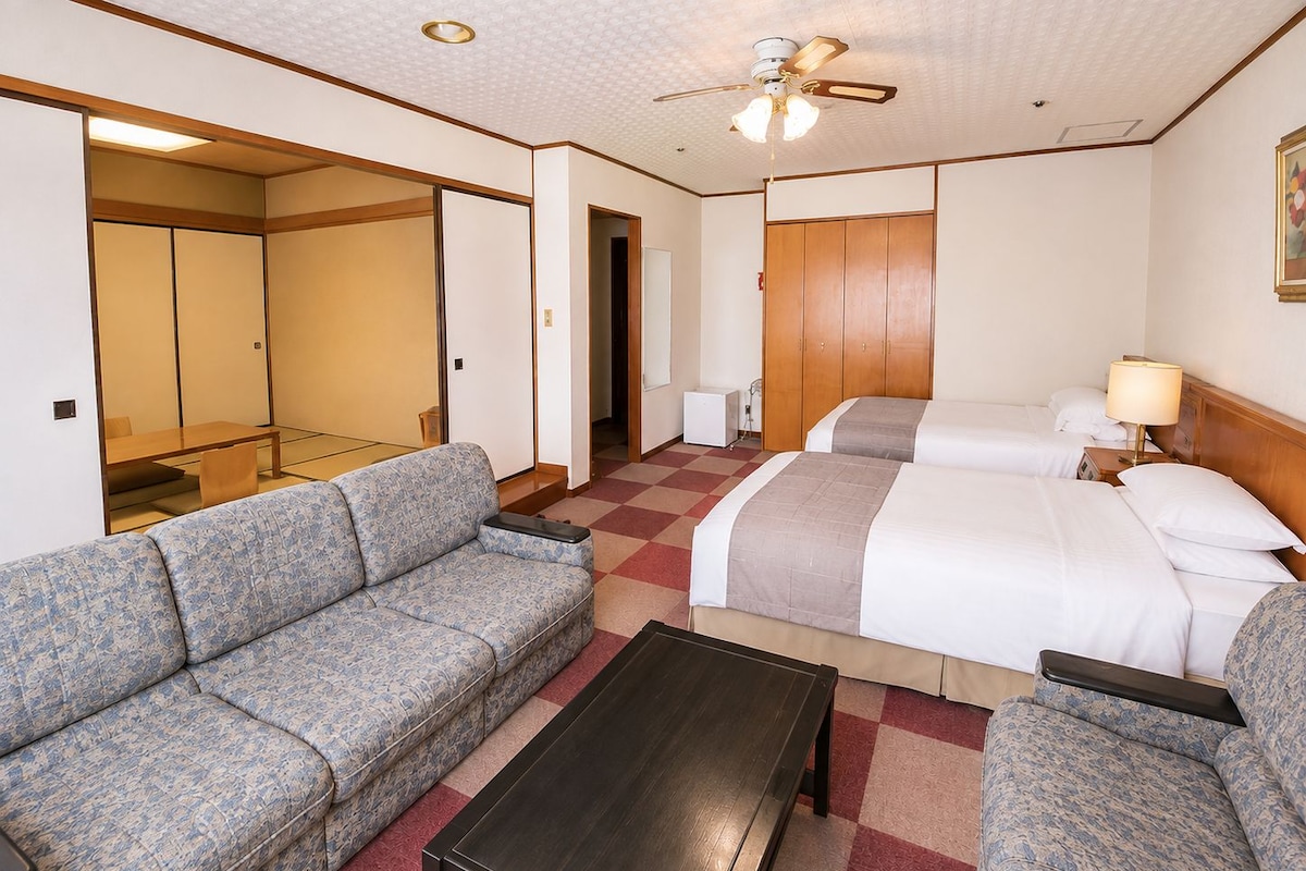 A hotel room with a sofa, two beds, a small table, and a ceiling fan.
