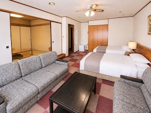 A hotel room with a sofa, two beds, a small table, and a ceiling fan. - New Sunpia Saitama Ogose (Ogose)