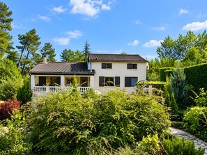 A two-story house with a white exterior and dark shutters, surrounded by lush greenery and a clear blue sky.