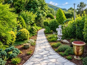 A stone pathway winding through a lush garden with various plants and a statue.