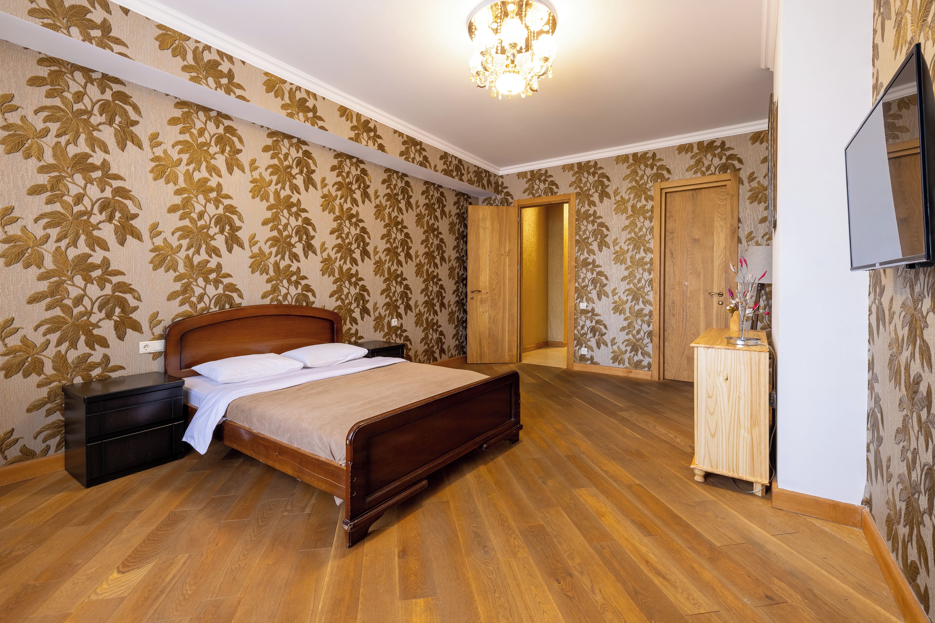 A bedroom with a wooden bed, a nightstand, a dresser, and a television on the wall.