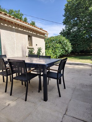 Une terrasse extérieure avec une table et des chaises, un bâtiment doté d’un toit en tuiles et une haie verte en arrière-plan.