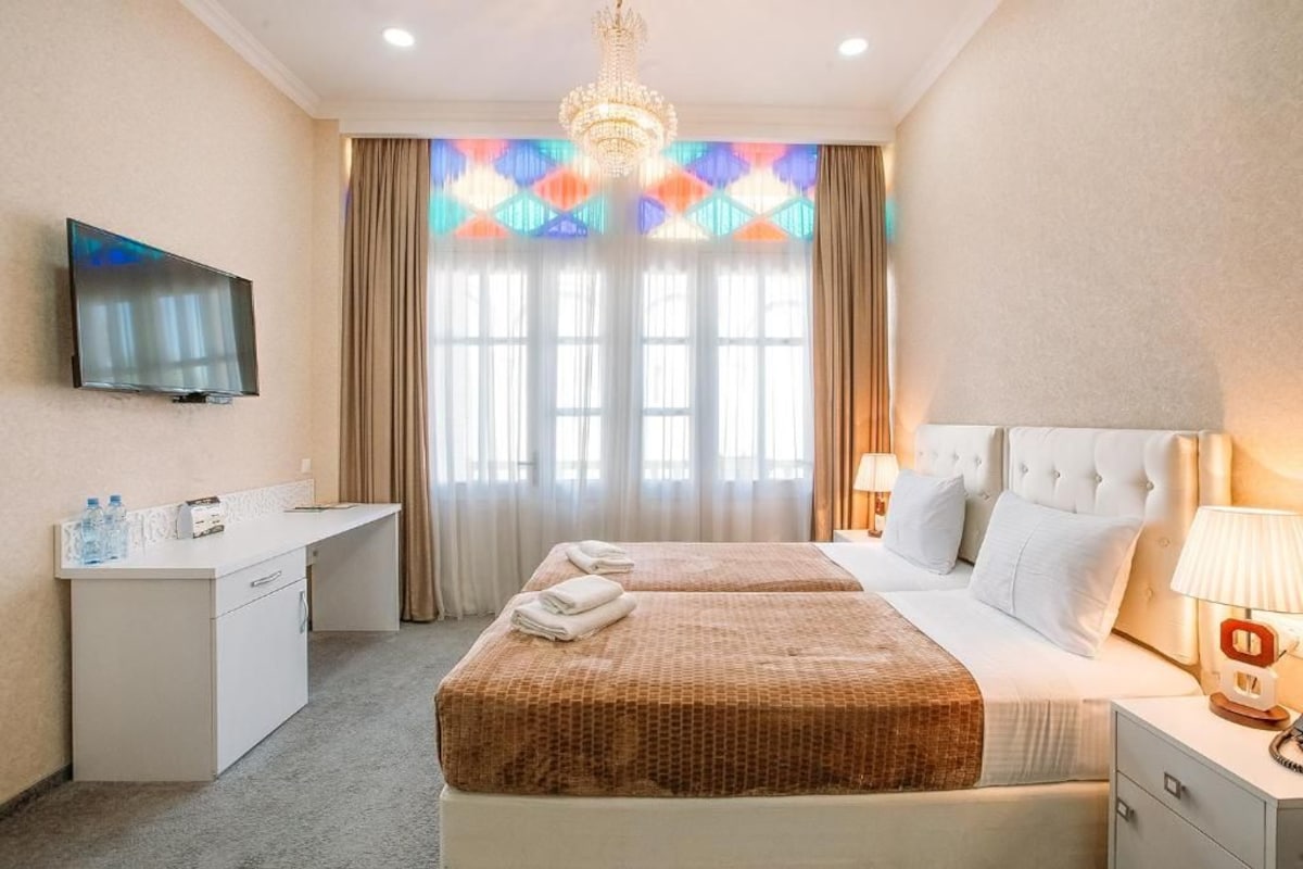 Quarto de hotel com cama, mesa de cabeceira, televisão, escrivaninha e cortinas.