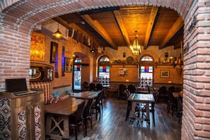 Restaurante com paredes de tijolo, vigas de madeira e janelas de vitrais. Mesas e cadeiras dispostas para refeições.