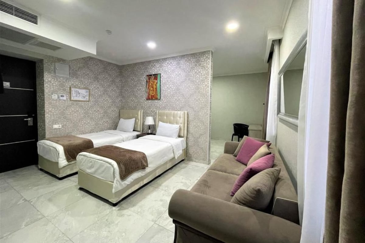 Quarto de hotel com duas camas de solteiro, um sofá, uma mesa de cabeceira com abajur e uma janela com cortinas.