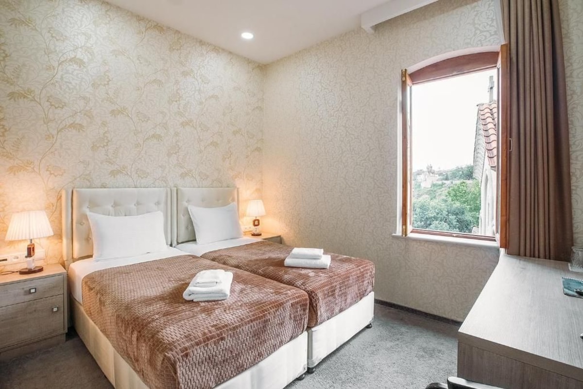 Quarto de hotel com cama, mesas de cabeceira, escrivaninha e janela com cortinas.