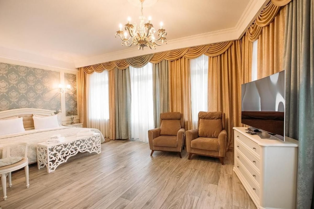 Quarto de hotel com cama, duas poltronas, televisão, penteadeira e lustre.
