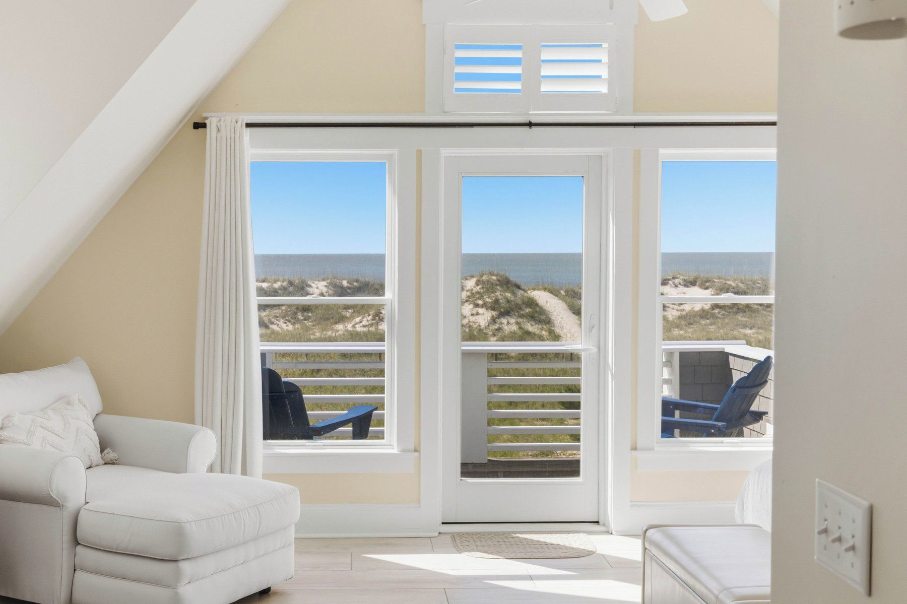 A room with a white sofa, a chair, and a view of the beach through a large window with shutters.