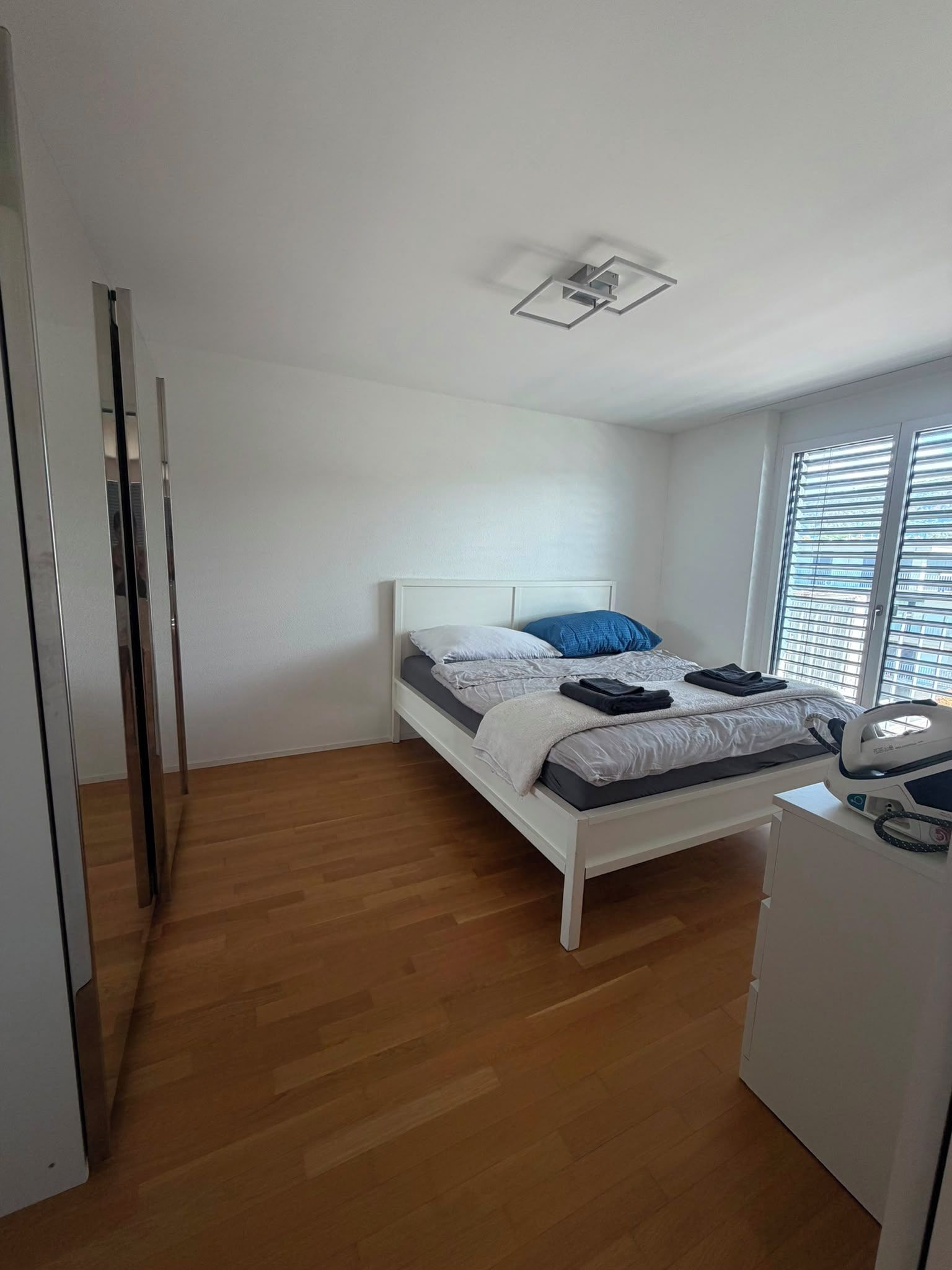 Um quarto com uma cama, janela com persianas, luminária no teto e piso de madeira.