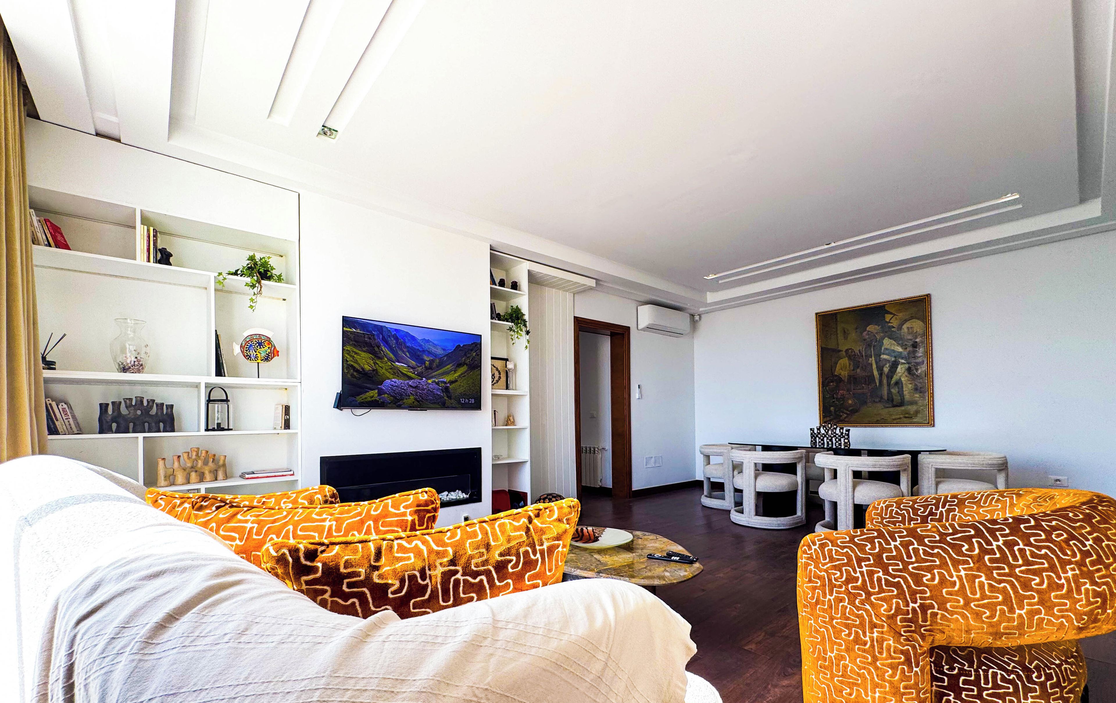 Ein modernes Wohnzimmer mit einer Couch, einem Couchtisch und einem an der Wand montierten Fernseher. Ein Bücherregal mit verschiedenen Gegenständen und ein Gemälde befinden sich über dem Fernseher.