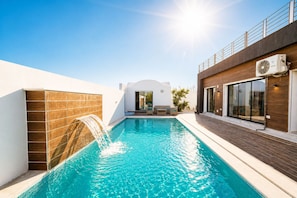 Una piscina con caída de agua, un edificio moderno con balcón y un cielo azul despejado.