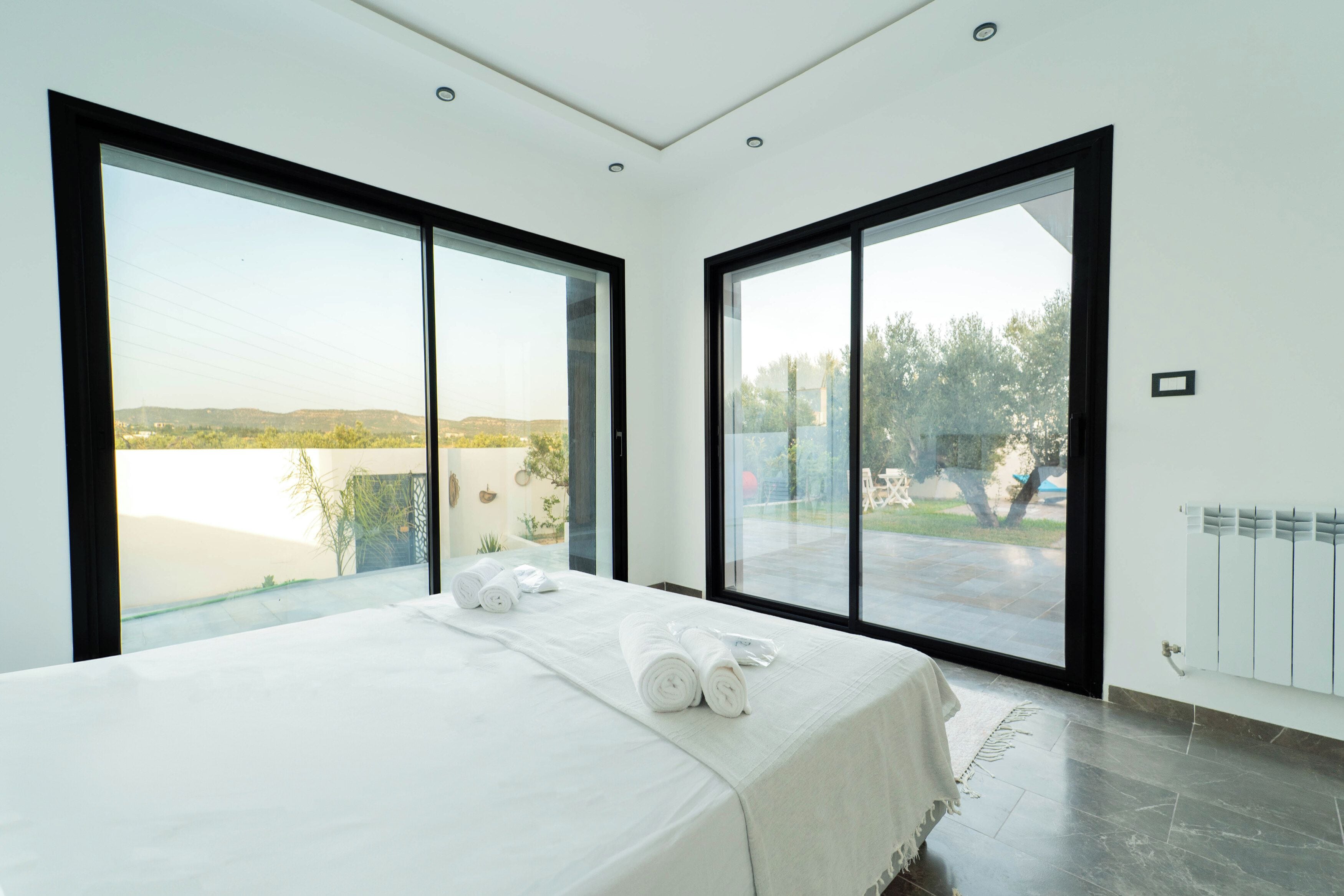 Un dormitorio moderno con una cama grande, dos ventanas y vista a un lago y colinas.