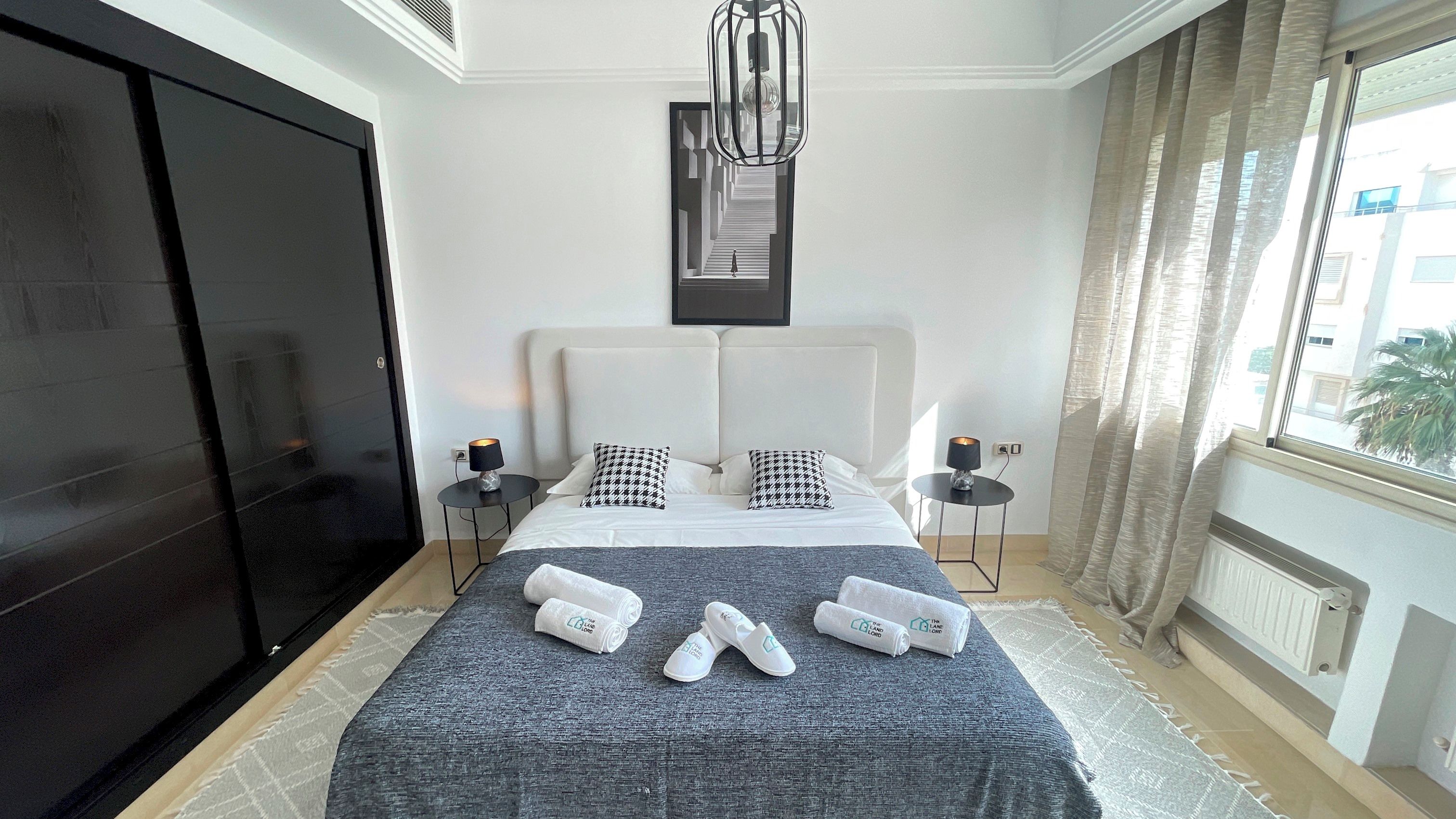 A hotel room with a bed, two bedside tables, a TV, a large window with curtains, and a radiator.