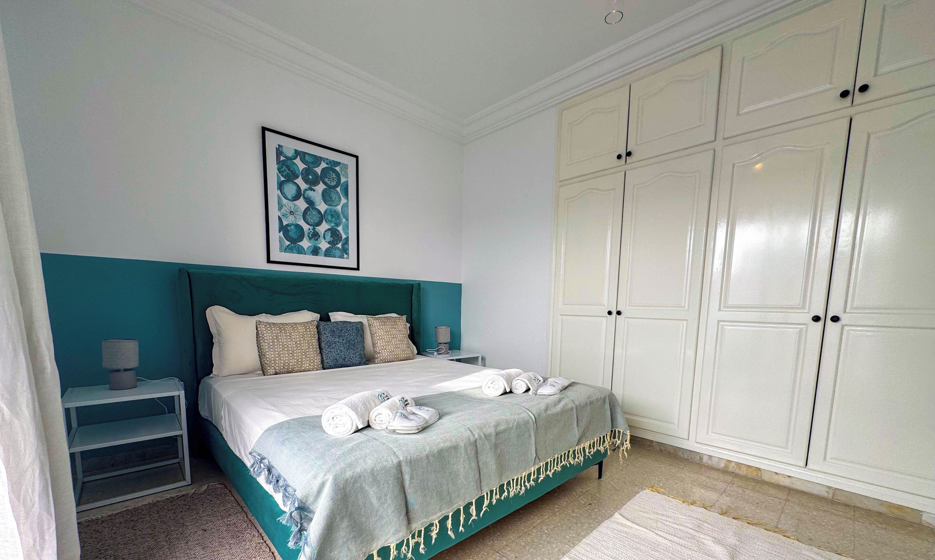 A neatly made bed with a blue headboard, a bedside table with a lamp, and a large white wardrobe.