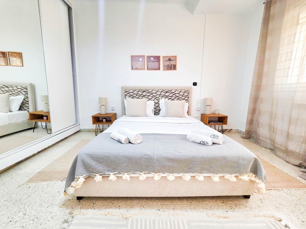 A neatly made bed with a grey bedspread and white pillows, a large mirror on one wall, and a small bedside table with a lamp.
