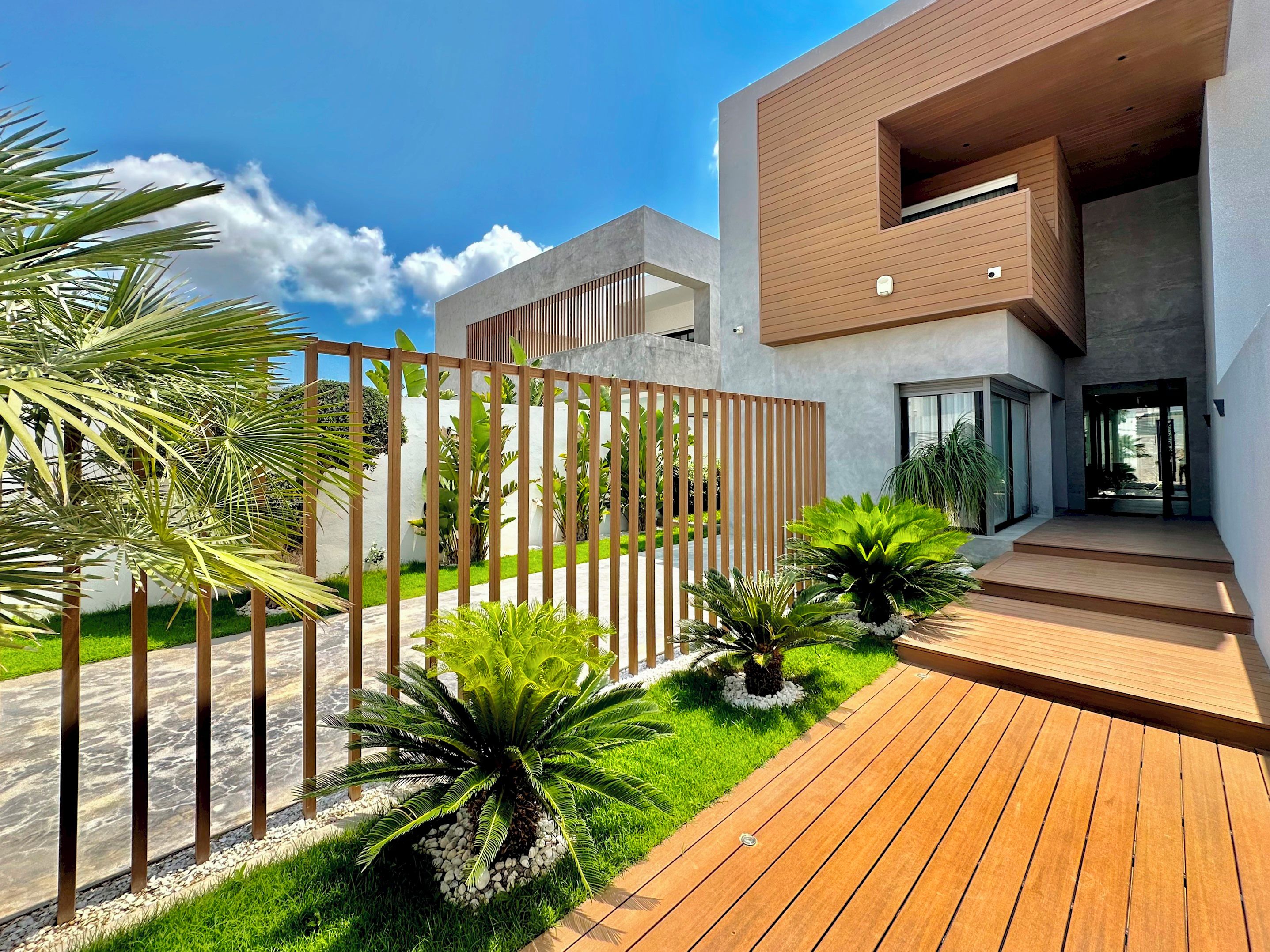 Un bâtiment moderne doté d’une terrasse en bois, d’une clôture en métal et d’une variété de plantes tropicales.