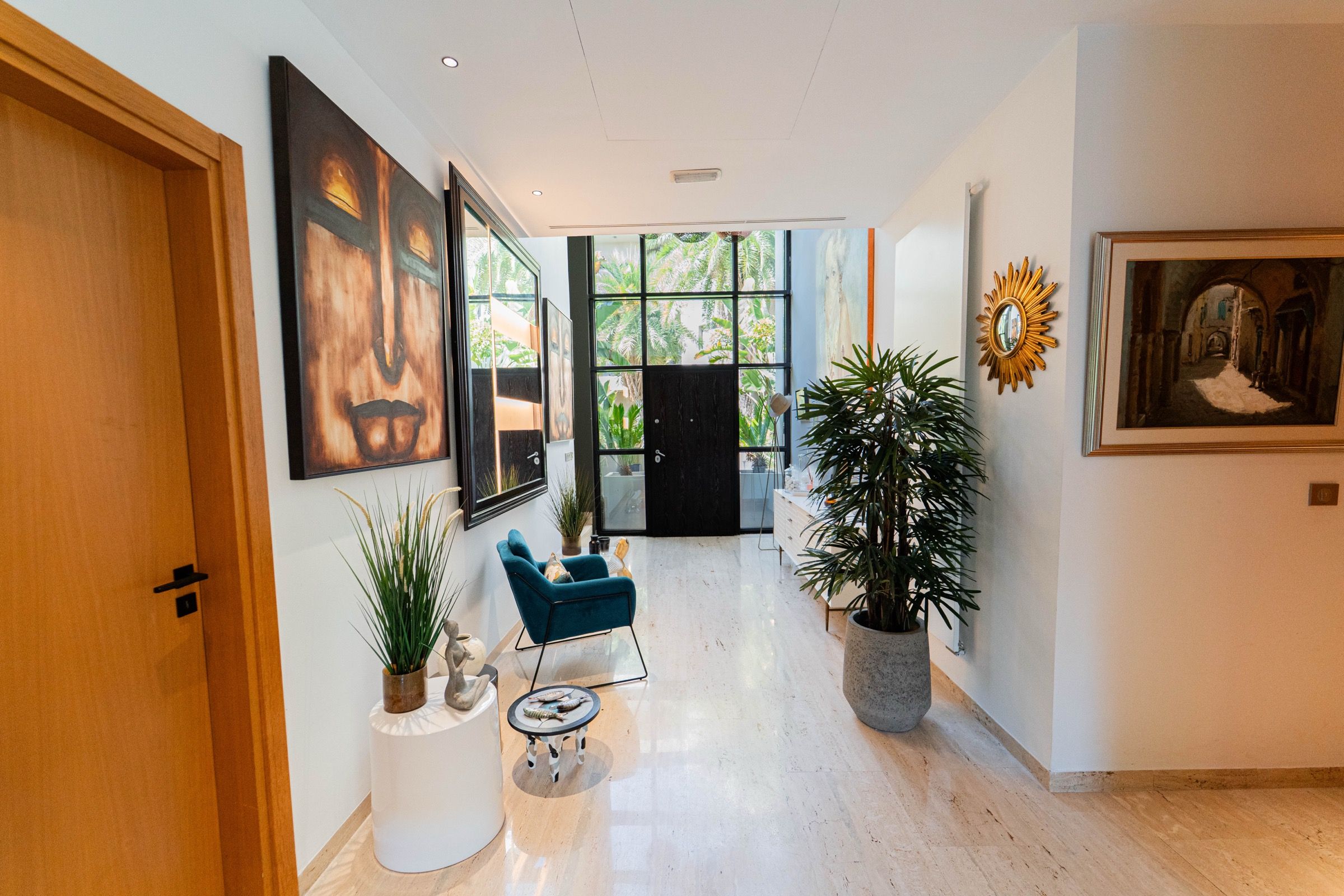 A hallway with a wooden door, a blue chair, a round table with fish, and a large potted plant.