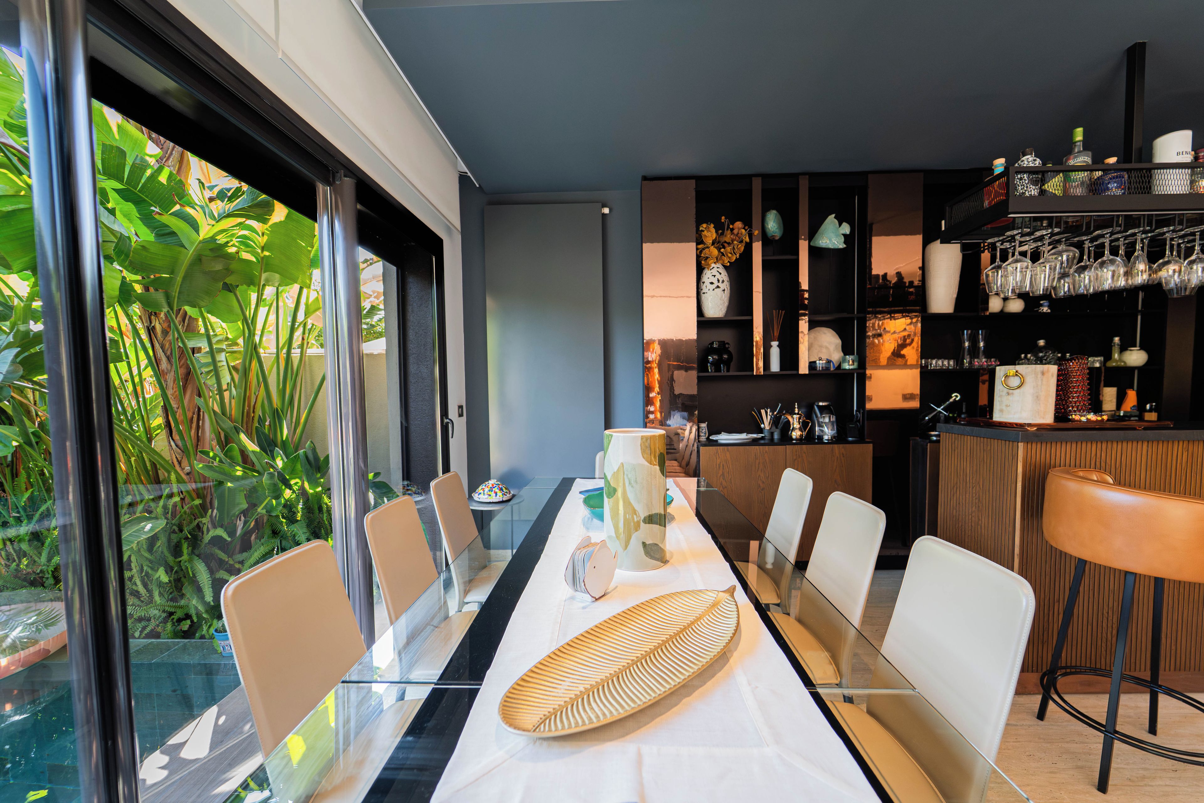 Un comedor moderno con una mesa de vidrio, sillas y una zona de bar con estantes y cristalería.