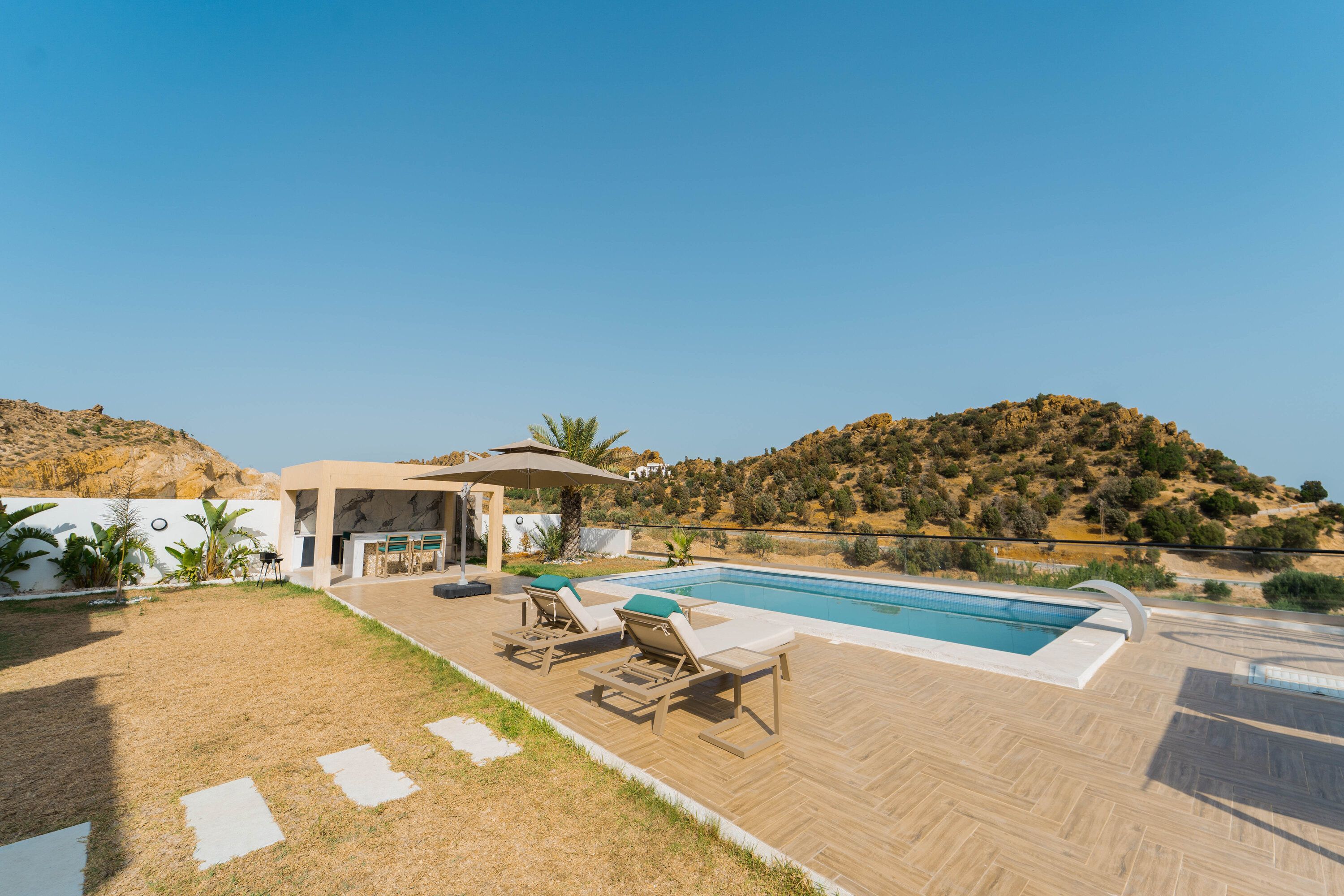 Zona de piscina con sillones, una barbacoa y vistas a un paisaje montañoso.