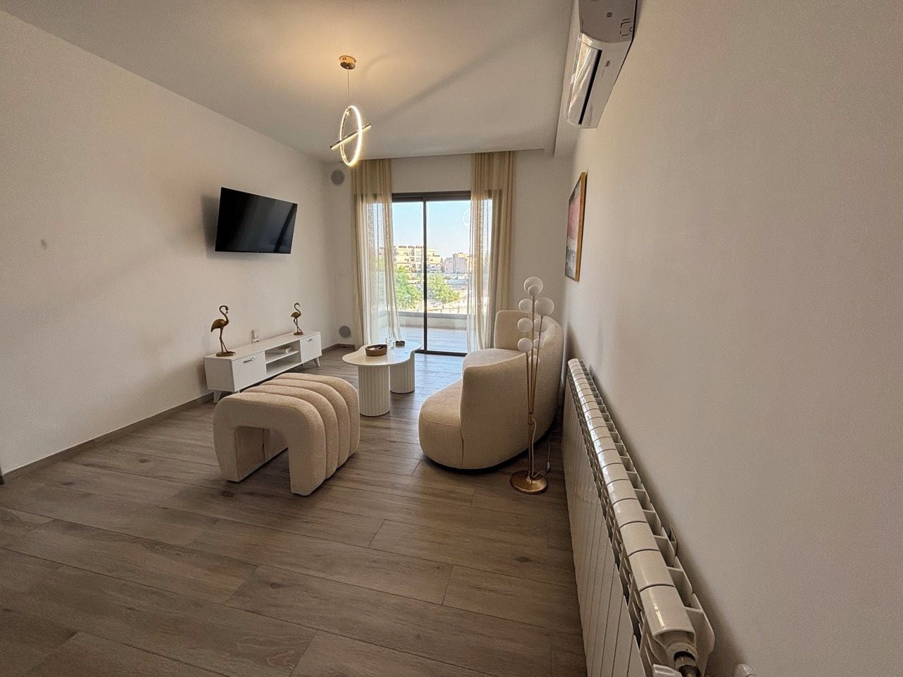 A modern living room with a flat-screen TV, a sofa, ottomans, a coffee table, and a floor lamp.