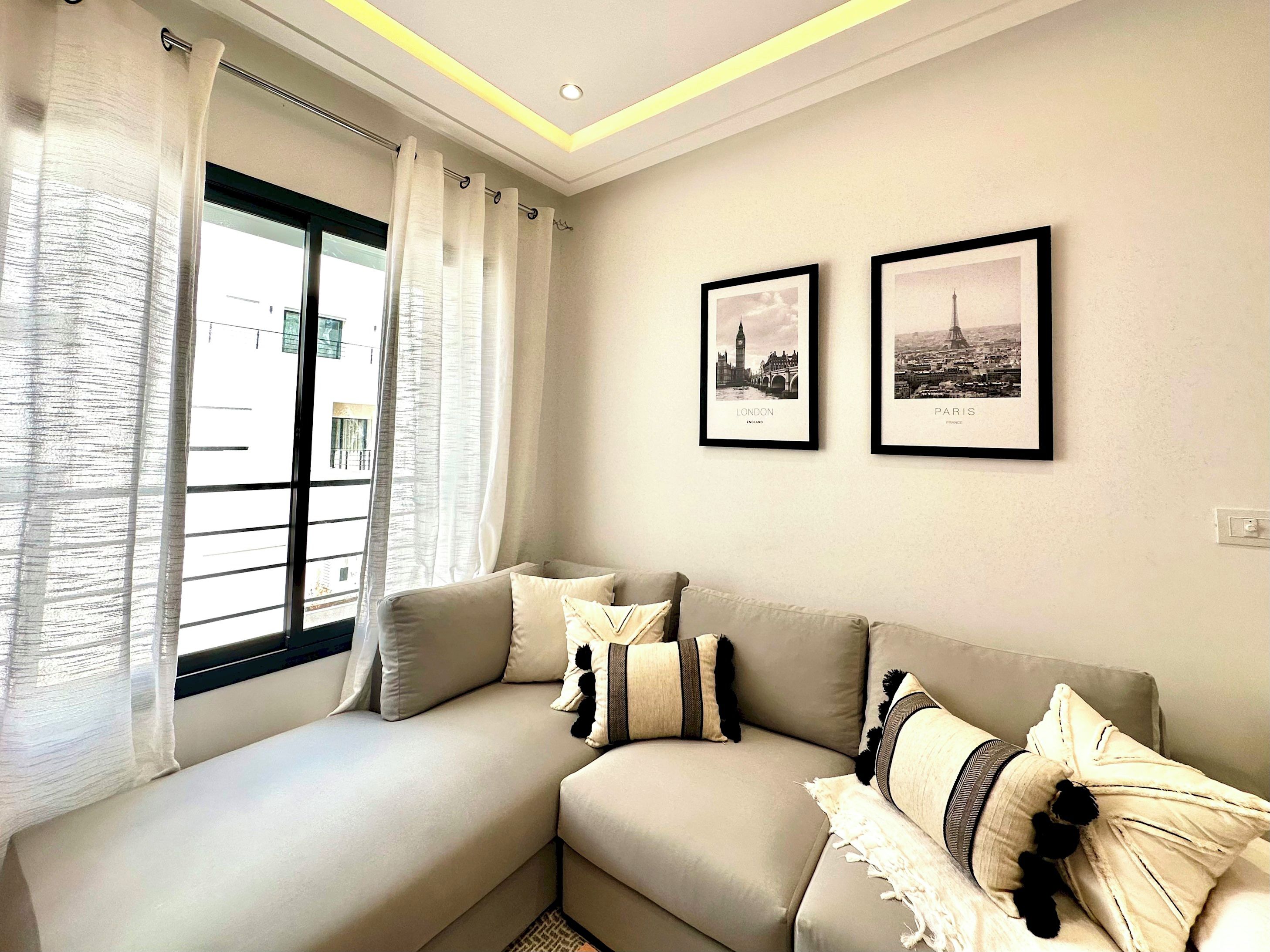 Una sala de estar moderna con un sofá, dos cuadros enmarcados de Londres y París, y un ventanal con cortinas.