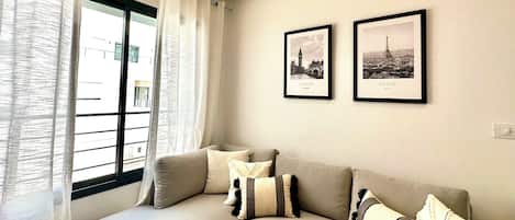Una sala de estar moderna con un sofá, dos cuadros enmarcados de Londres y París, y un ventanal con cortinas.
