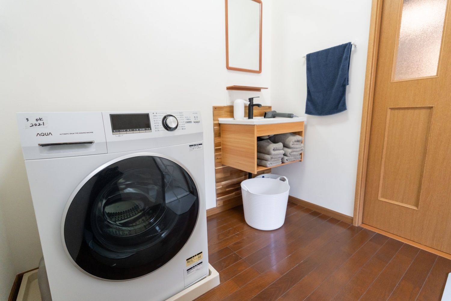 A compact, modern laundry room with a front-loading washing machine, a wooden shelf with folded towels, a white laundry basket, and a wooden door.