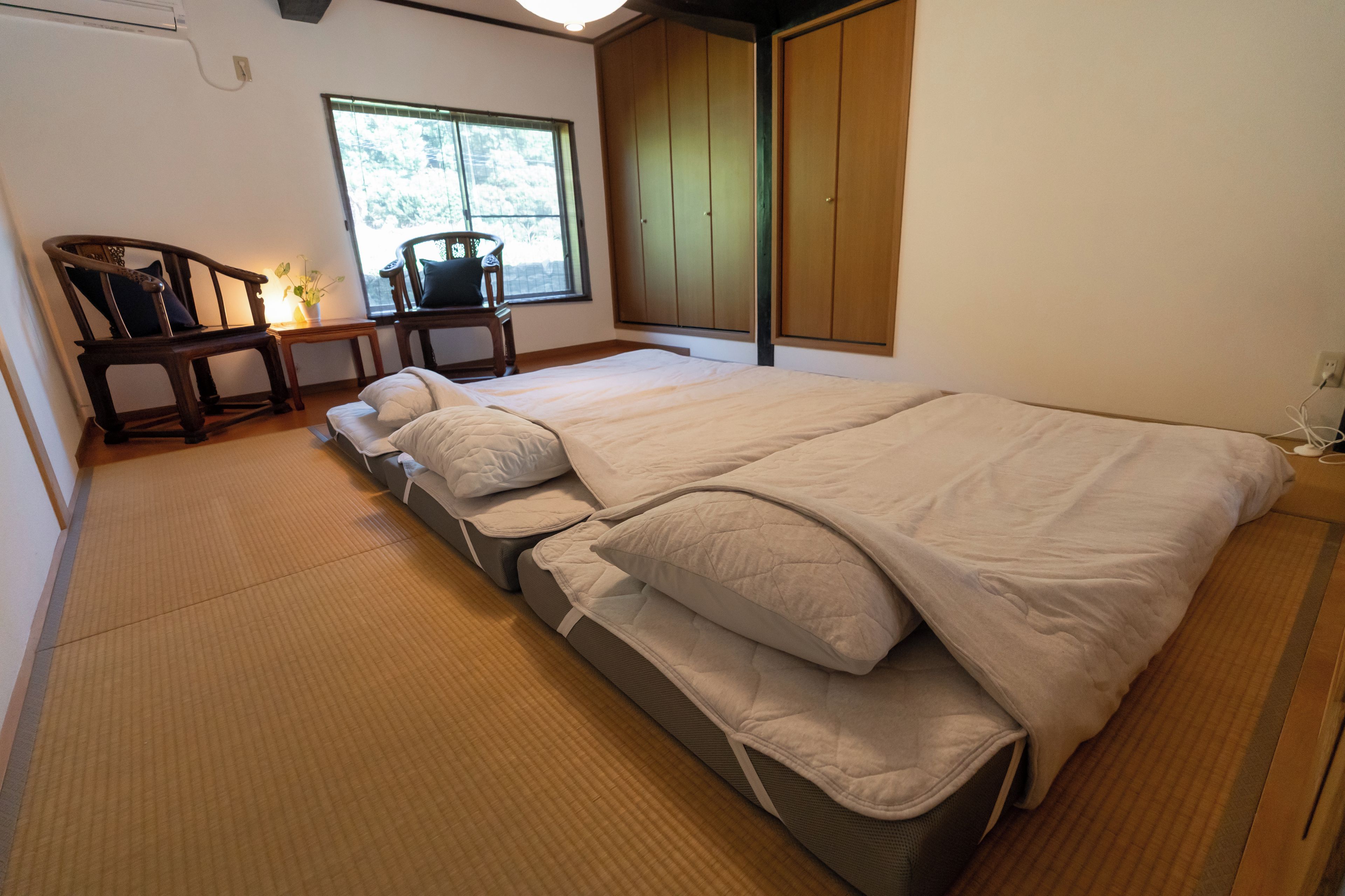 A traditional Japanese-style bedroom with tatami flooring, a low bed with multiple pillows, a wooden desk with a chair, and a wardrobe.