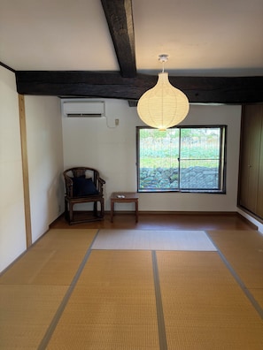 A room with tatami flooring, a traditional Japanese air conditioning unit, a wooden chair, a small table, and a large window with a view of greenery.