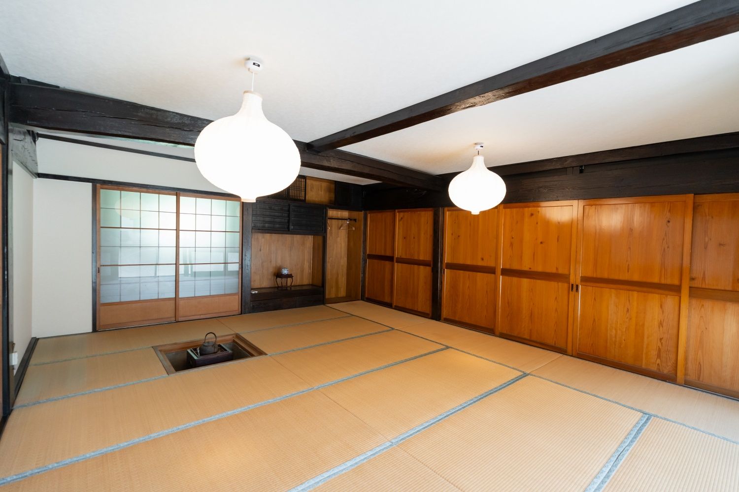 Um espaço com tatames, portas corrediças, uma mesa pequena com uma chaleira e duas luminárias suspensas.