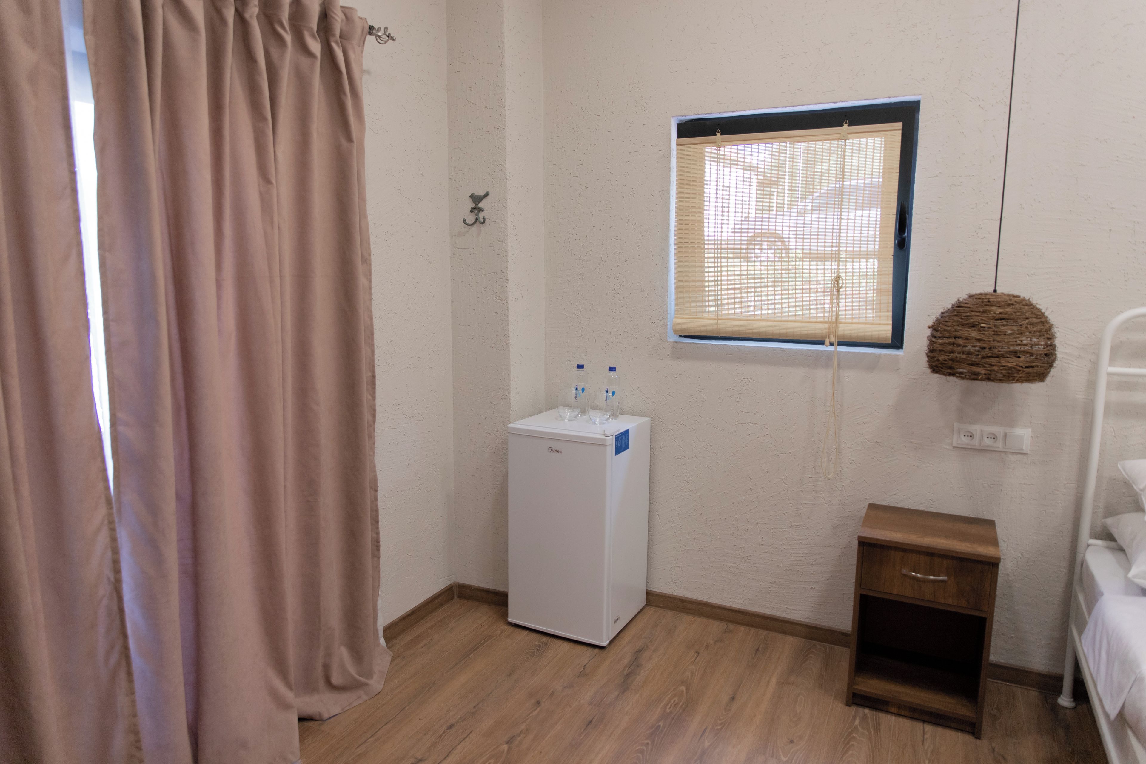 Una habitación pequeña y limpia con un refrigerador blanco, una mesita de noche, un cesto colgante y una ventana con persianas.