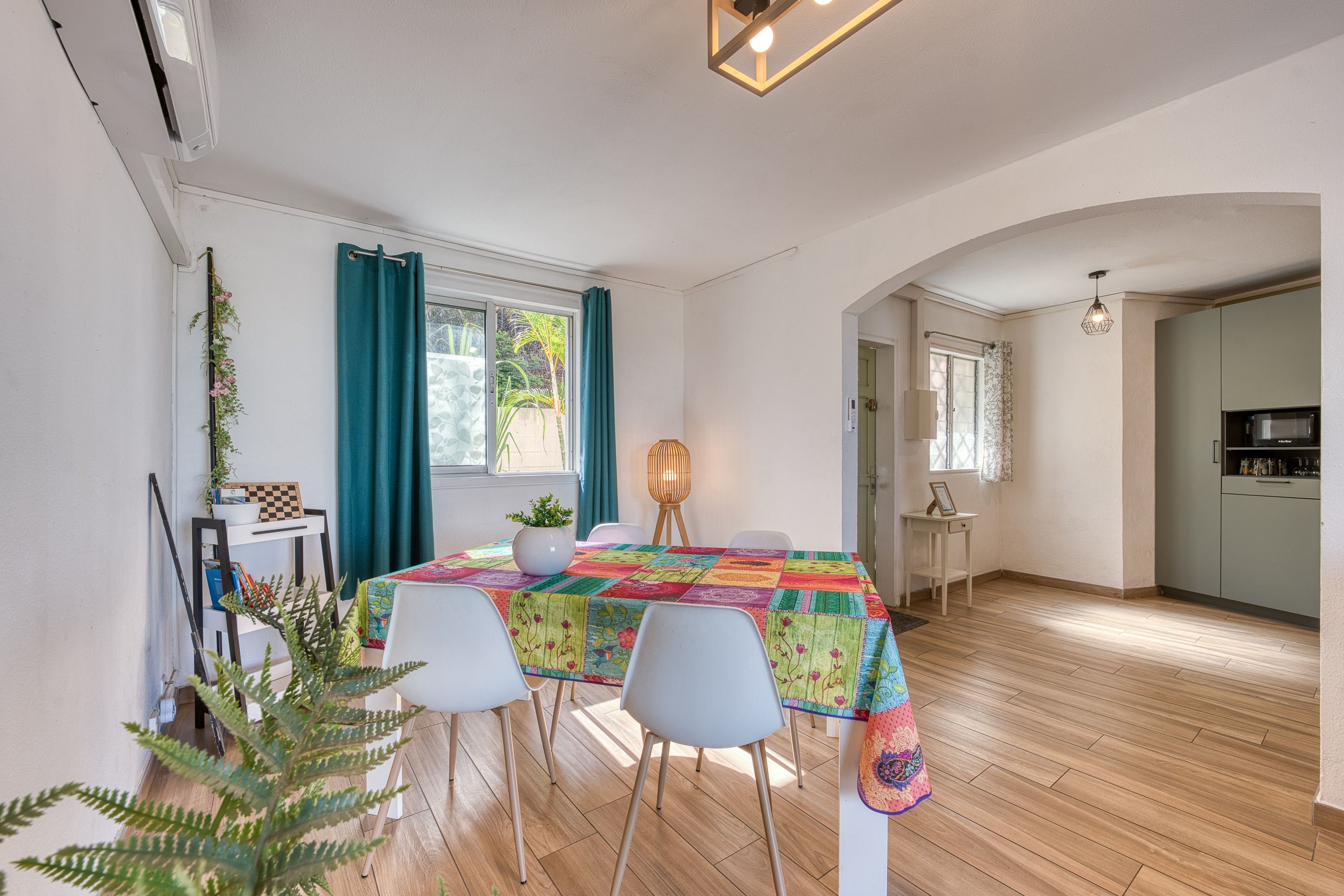 A dining area with a table covered by a colorful quilt, white chairs, a potted plant, and a window with curtains.